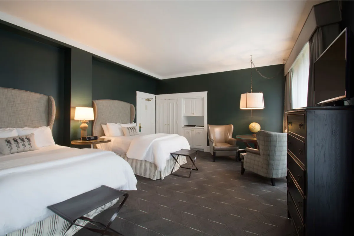 The image showcases a spacious, sunlit hotel room with two plush beds featuring elegant headboards, a stylish armchair perfect for reading by the window, a modern lamp, and a sleek dresser. The overall mood is restful and inviting, with gentle morning light filling the space, suggesting comfort and relaxation after exploring the city.