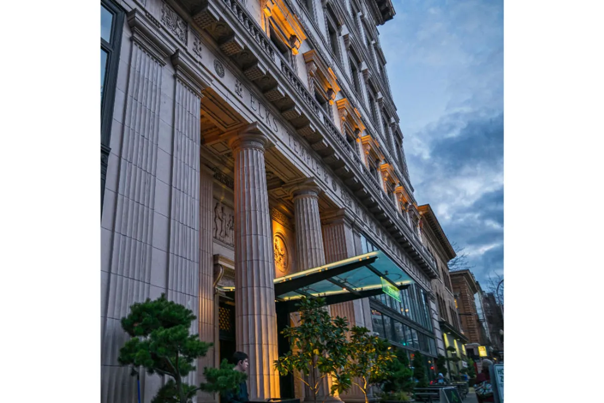 An exterior view of the hotel at dusk, showcasing illuminated ornate columns, a glass entrance canopy, and lush potted plants. The scene feels lively and welcoming, with hints that people are gathering or socializing outside, suggesting activity and connectivity with the city.