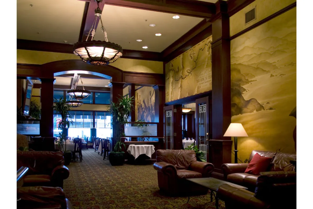 An elegantly appointed lounge featuring ornate chandeliers, lush greenery, comfortable leather sofas, and a detailed wall mural. The space is welcoming and sophisticated, ideal for both casual and intimate gatherings.