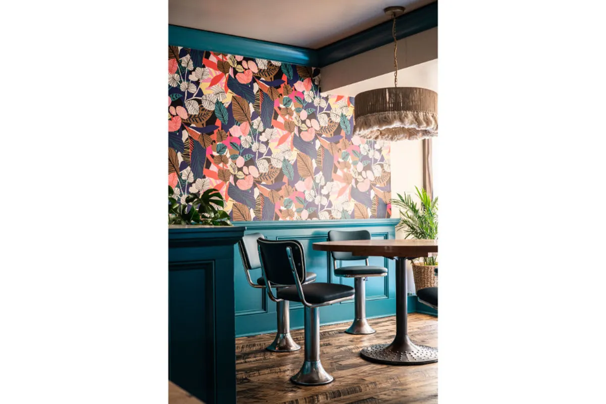 A stylish, light-filled dining nook featuring modern chairs gathered around a round table, set against a backdrop of bold floral wallpaper and an elegant pendant light. Sunlight streams through nearby windows, enhancing the cheerful and relaxed ambiance.