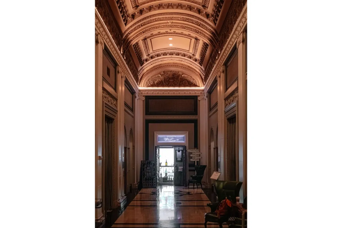 A grand hallway with intricately detailed ceilings, an elegantly patterned floor, and a majestic entrance at the end. Ornate wall decorations add to the regal atmosphere, and the spacious area is illuminated by soft, natural light.