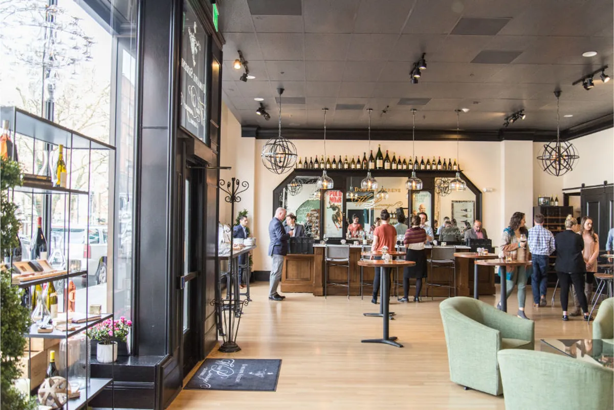 A lively bar interior with a polished counter lined with wine bottles and guests engaging in relaxed conversation. Comfortable seating surrounds the area, creating a sociable and inviting ambiance. The lighting is warm and the mood encourages mingling and enjoyment of drinks.