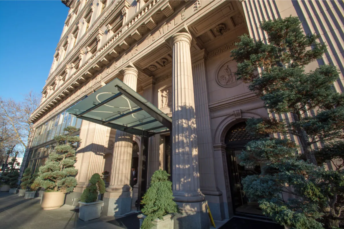 The image captures the hotel's stately exterior entrance, featuring grand columns, an elegant glass canopy, and well-manicured trees flanking the approach. The inviting outdoor area suggests opportunities for guests to socialize or take in the architectural details while stepping out into the surrounding neighborhood.