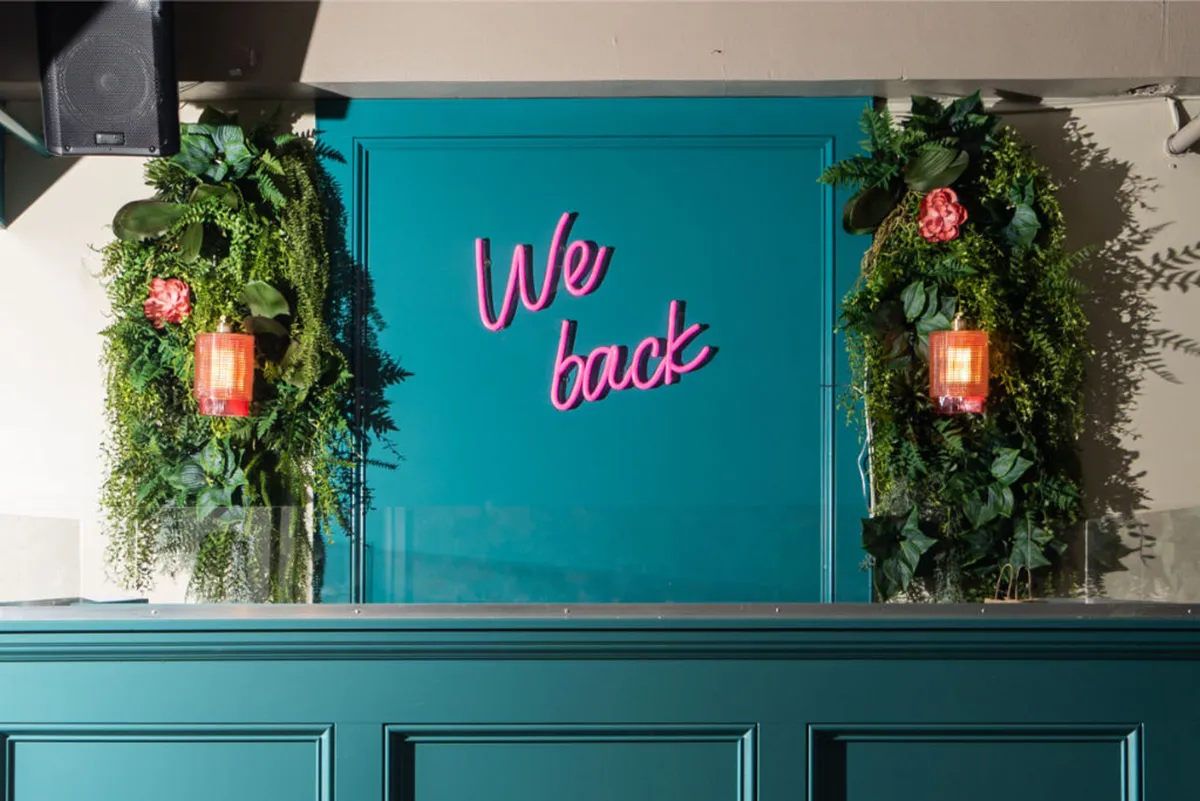 A lively interior space featuring a neon sign reading 'We back,' lush green foliage, and bright orange lanterns. The area is arranged for casual gatherings and offers a welcoming, communal feel that invites guests to relax or socialize after a day of city exploration.